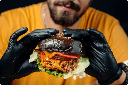 Hands in black gloves holding up a burger on a dark bun, stacked with cheese, tomato and lettuce